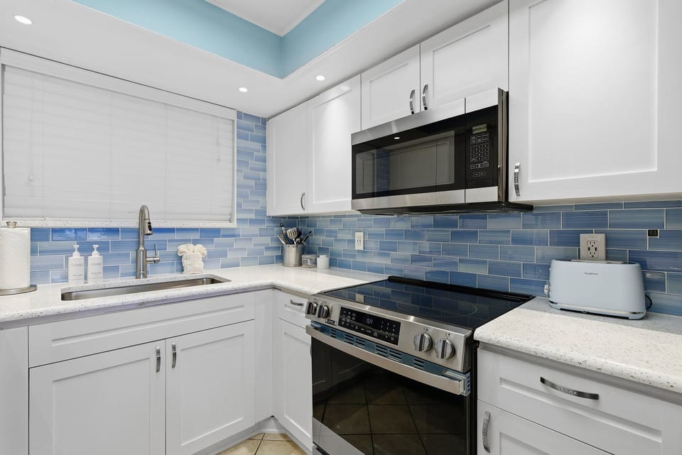 Cook with ease in this stylish coastal kitchen featuring modern appliances, bright white cabinetry, and blue tile backsplash. A perfect addition to your beach stay, offering a fresh and functional space after a day in the sun.