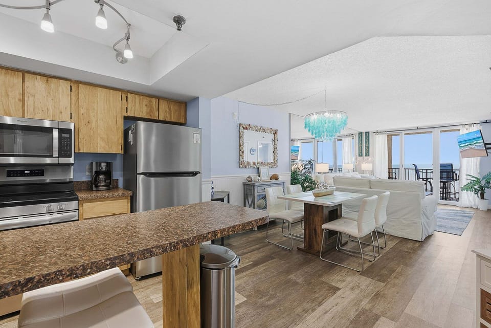 Bright open-concept layout featuring a full kitchen, dining area, and living space with ocean views. Perfect for a beach stay, enjoy seamless indoor-outdoor living just steps from the balcony and shoreline.