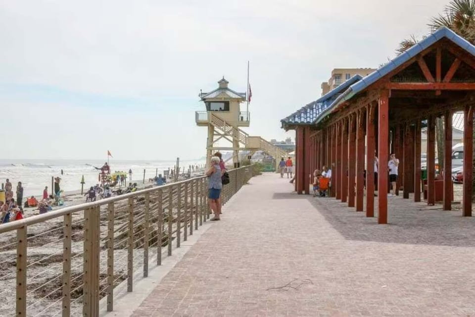 A serene coastal walkway next to the beach, featuring a lifeguard tower, shaded seating, and ocean views. Perfect for relaxing and enjoying the lively beach atmosphere.