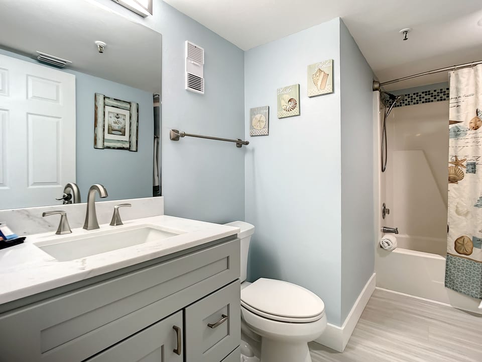 This clean, beach-inspired bathroom features a spacious vanity with modern fixtures, a cozy bathtub, and coastal decor. The inviting atmosphere provides a refreshing space for guests to relax and rejuvenate after a day at the beach.