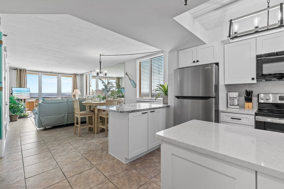 Enjoy ocean views from this modern open-concept kitchen and living space, featuring sleek white cabinetry, stainless steel appliances, and a cozy dining area perfect for gatherings by the sea.