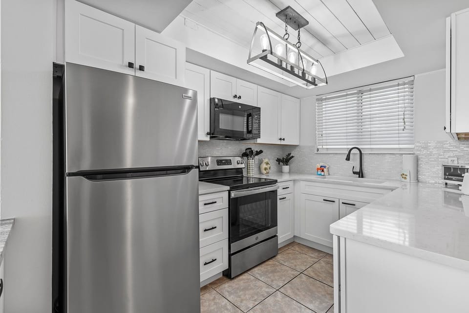 This sleek kitchen boasts stainless steel appliances, white cabinetry, and a stylish light fixture. The black accents and large window add a touch of sophistication to this functional and well-designed cooking space.