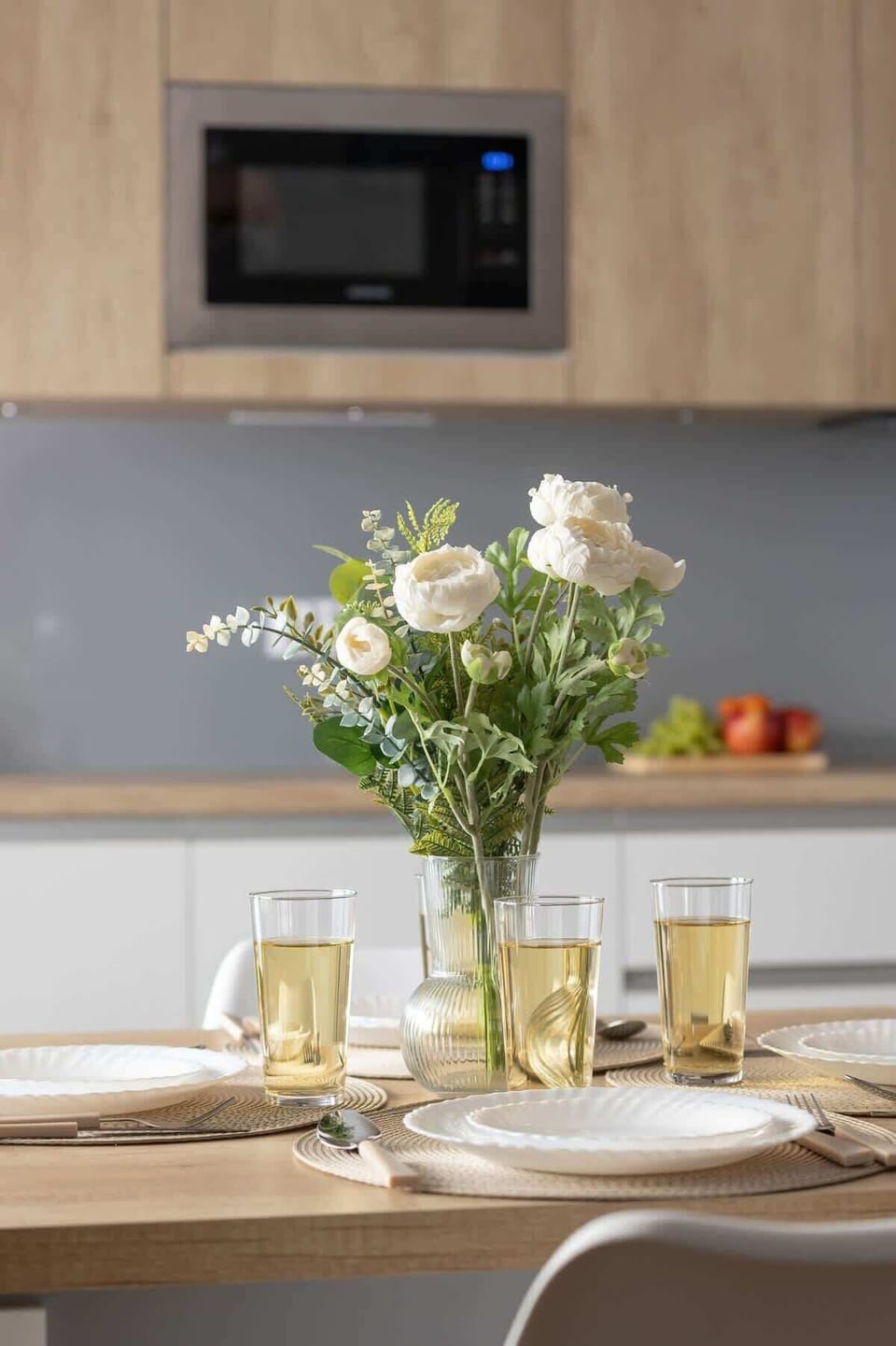 A stylish kitchen area with wooden cabinets, a built-in microwave, and a dining table with a vase of fresh flowers, creating a warm and inviting atmosphere.

