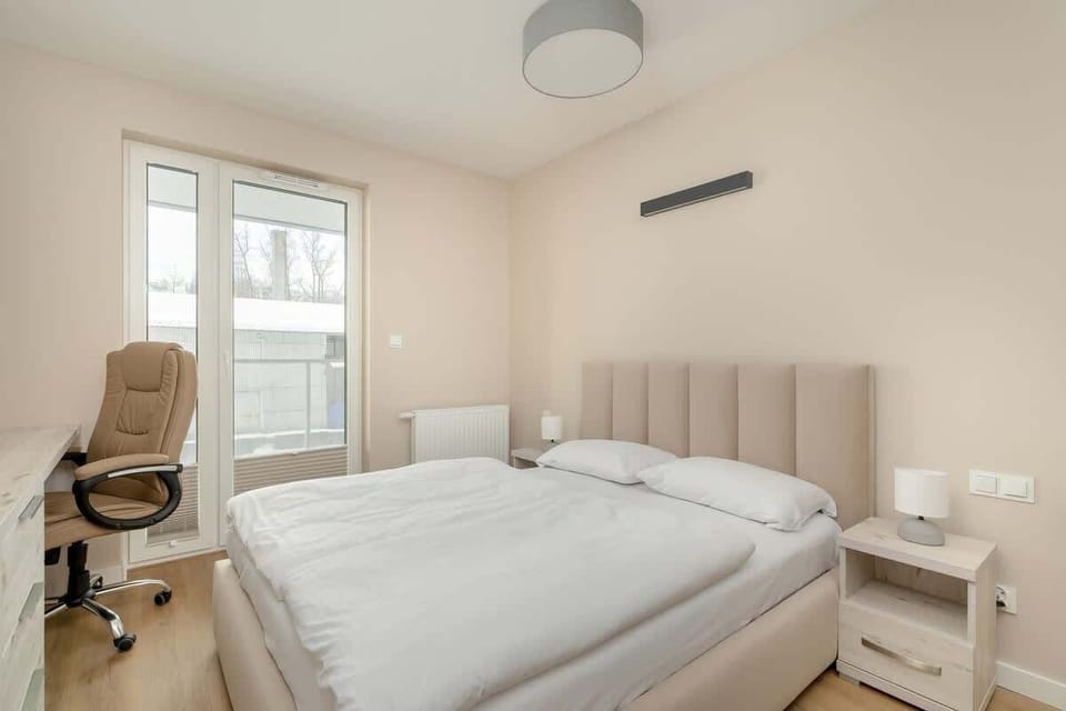 A cozy bedroom featuring a double bed with a neutral headboard, a bedside table, and a soft overhead light. A window provides a pleasant outdoor view.

