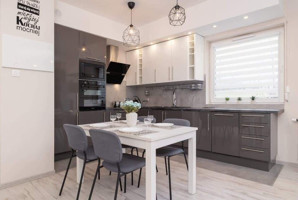A contemporary kitchen and dining area with dark cabinets, a white countertop, and a dining table with four elegant chairs, illuminated by hanging light fixtures.

