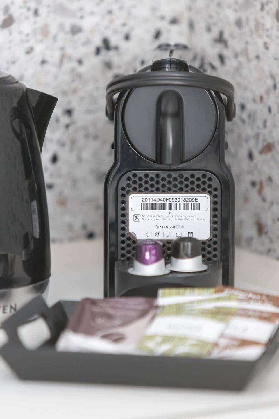 A close-up of a compact coffee machine with neatly arranged coffee pods, set against a terrazzo-patterned background.
