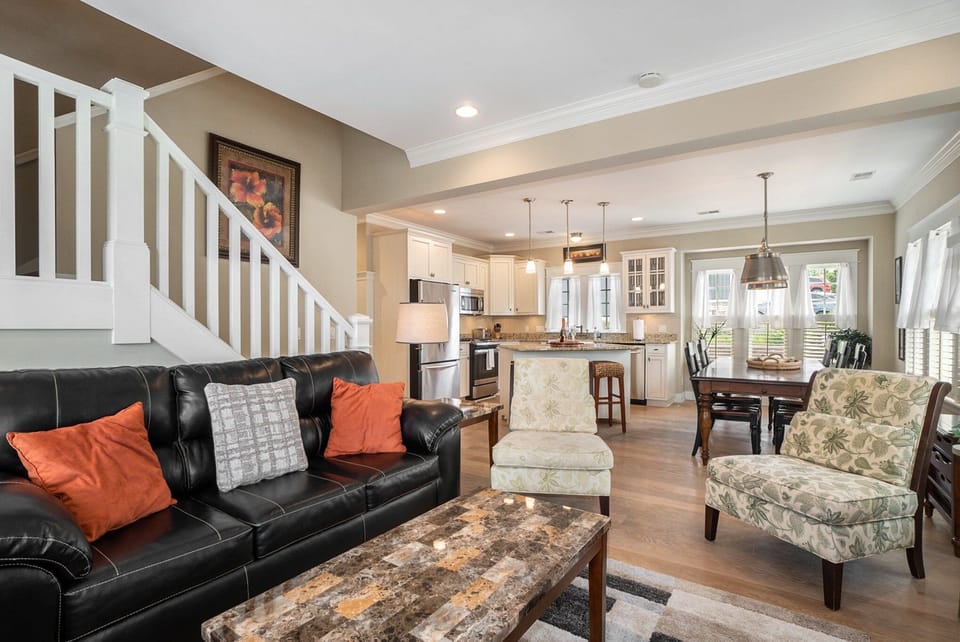 Features a black leather sofa with orange and grey throw pillows, a marble-topped coffee table, and two patterned armchairs. The space seamlessly transitions into a kitchen and dining area.