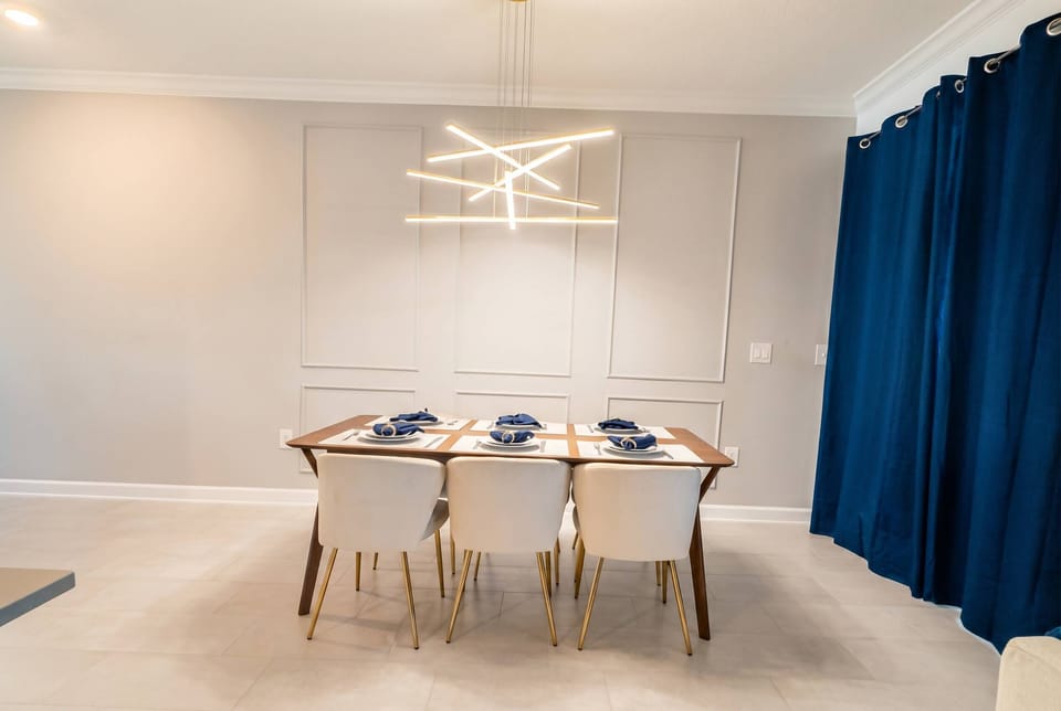 Experience a stylish dining area featuring a modern light that gently illuminates a table for four, with crisp white chairs against soft gray walls, complemented by elegant dark curtains for a chic ambiance.