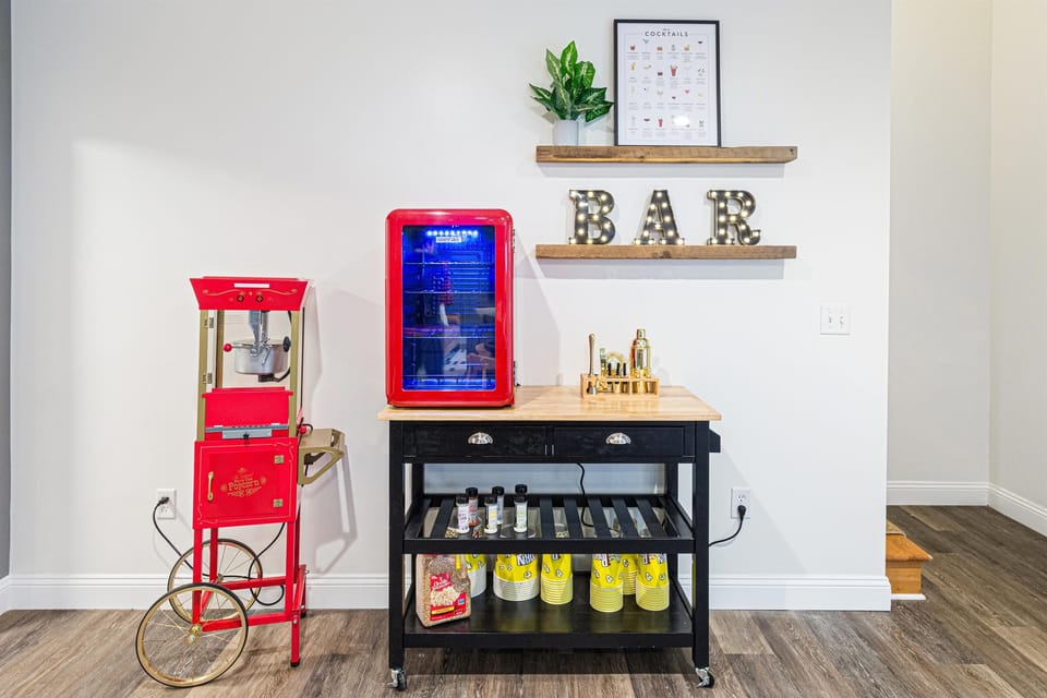 The bar area on the lower level — vintage-style popcorn machine (popcorn and seasonings provided), mini fridge for drinks, cocktail guide on the wall, and a light-up BAR sign.