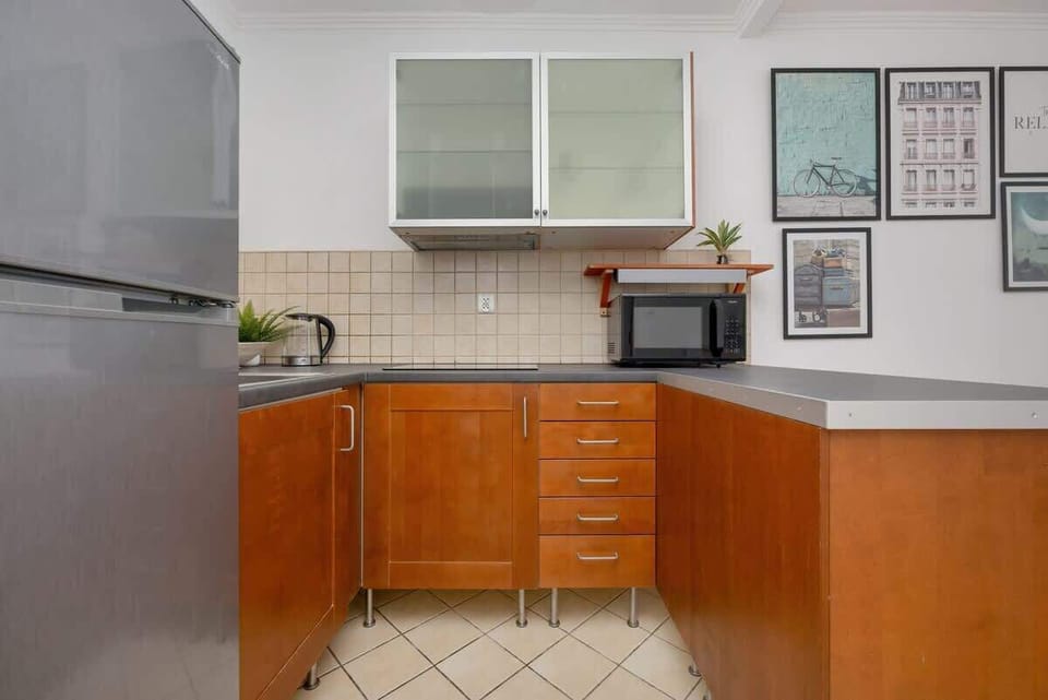 A compact kitchen featuring wooden lower cabinets, white upper cabinets with frosted glass, a stainless steel refrigerator, and a microwave placed on the countertop. The space is well-lit and decorated with framed artwork.

