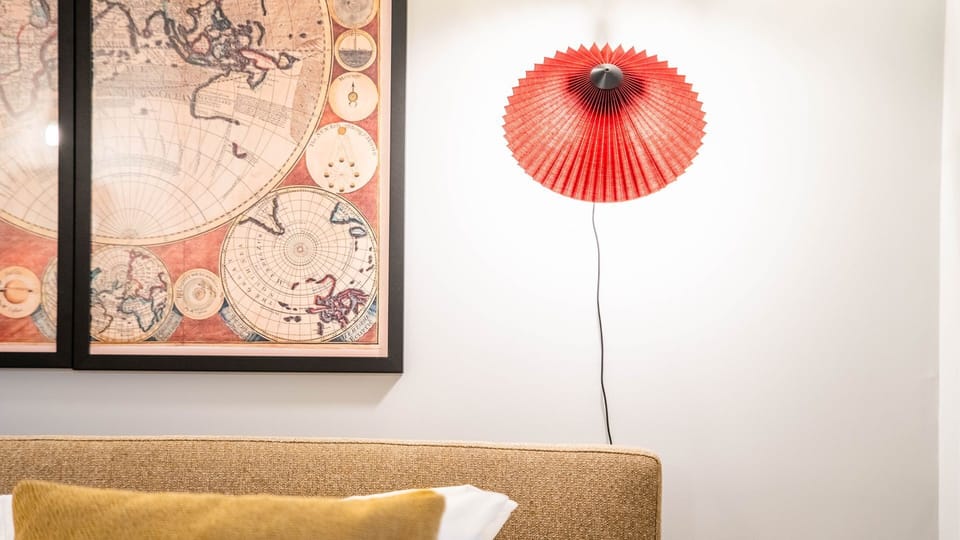 Drift off to sleep in a uniquely styled bedroom, featuring a comfortable headboard complemented by a captivating framed map artwork and a distinctive red pleated wall lamp. This cozy nook invites relaxation and adds a touch of artistic flair to your priva