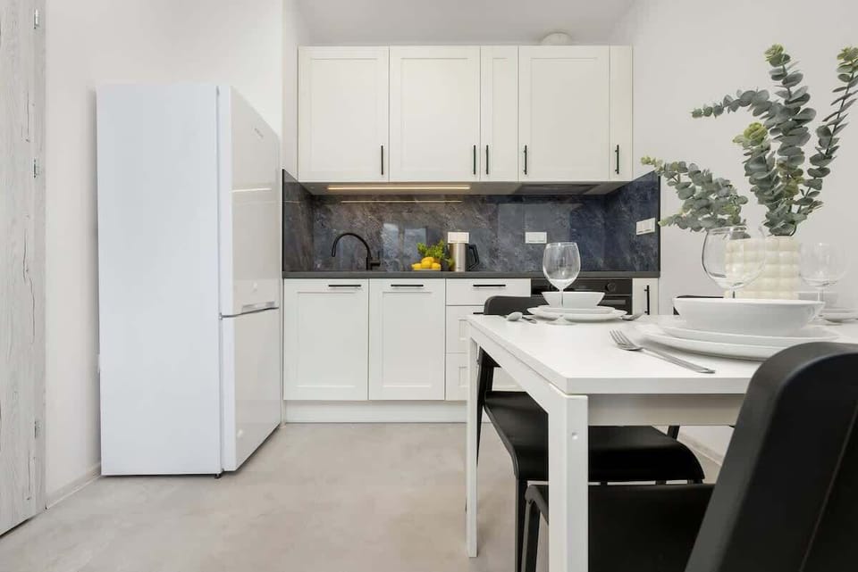 A different perspective of the kitchen, featuring a full-sized refrigerator, white cabinets, and a small dining setup with a plant centerpiece.

