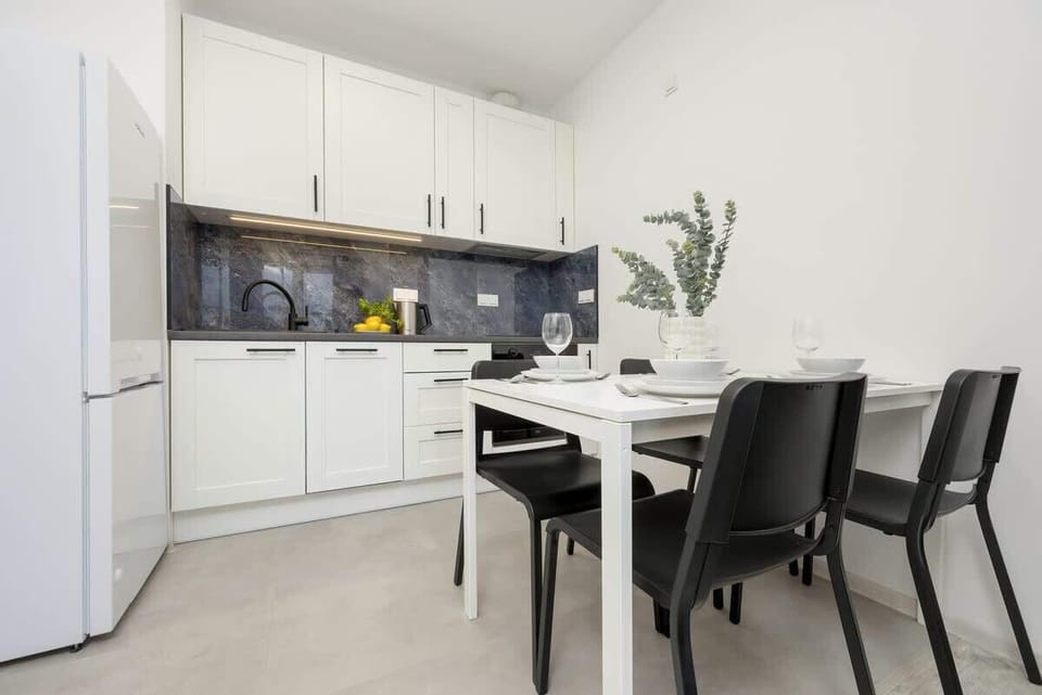 A compact yet stylish kitchen with white cabinetry, black countertops, and a dining table with two chairs, accompanied by a small plant for decoration.

