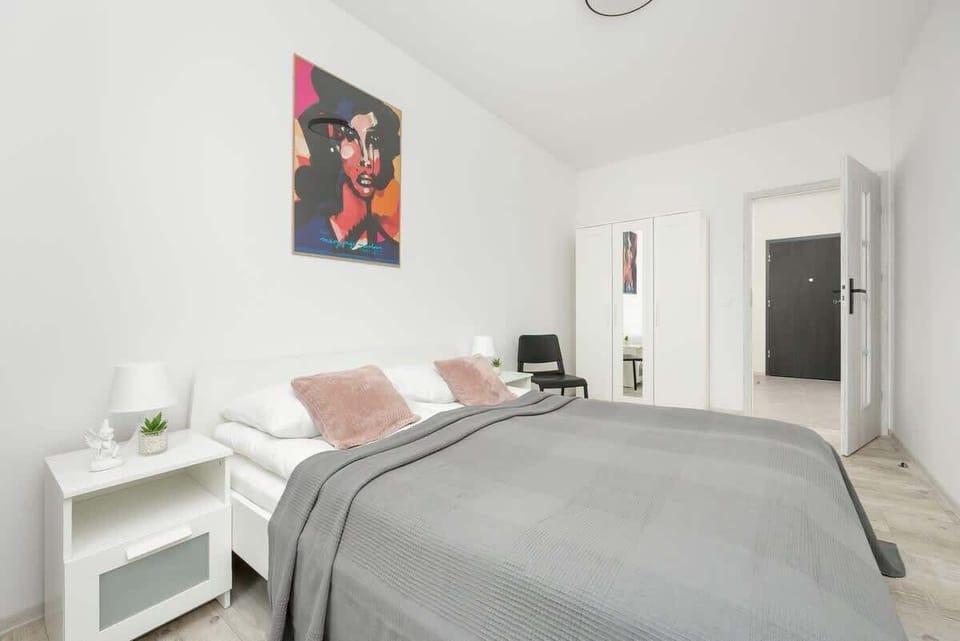 A cozy bedroom with a neatly made bed covered in a gray duvet, complemented by soft pink accent pillows, a white bedside table with a lamp, and a stylish red painting on the wall.

