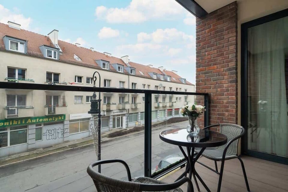 A private balcony with a glass railing, a black metal table with chairs, and a charming view of the surrounding residential neighborhood with red-roofed houses.
