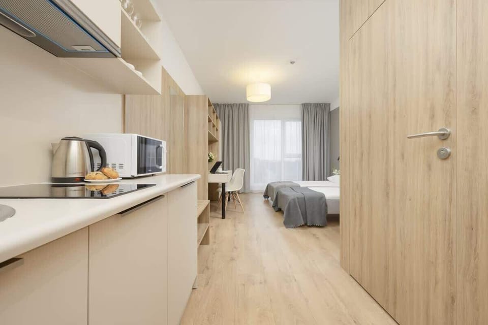 A sleek kitchenette with light wood and white accents, equipped with a kettle, storage cabinets, and a narrow layout maximizing space efficiency.

