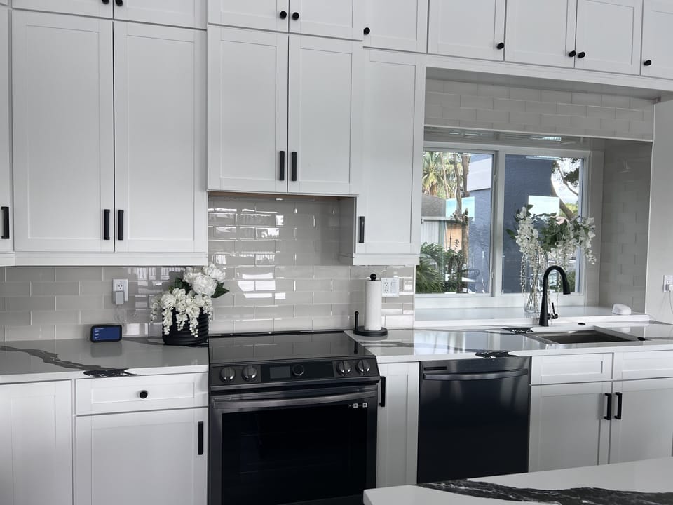 A modern kitchen with white cabinets, black appliances, a marble countertop, a black faucet, and a window above the sink. There is a vase with white flowers and a paper towel holder on the counter.
