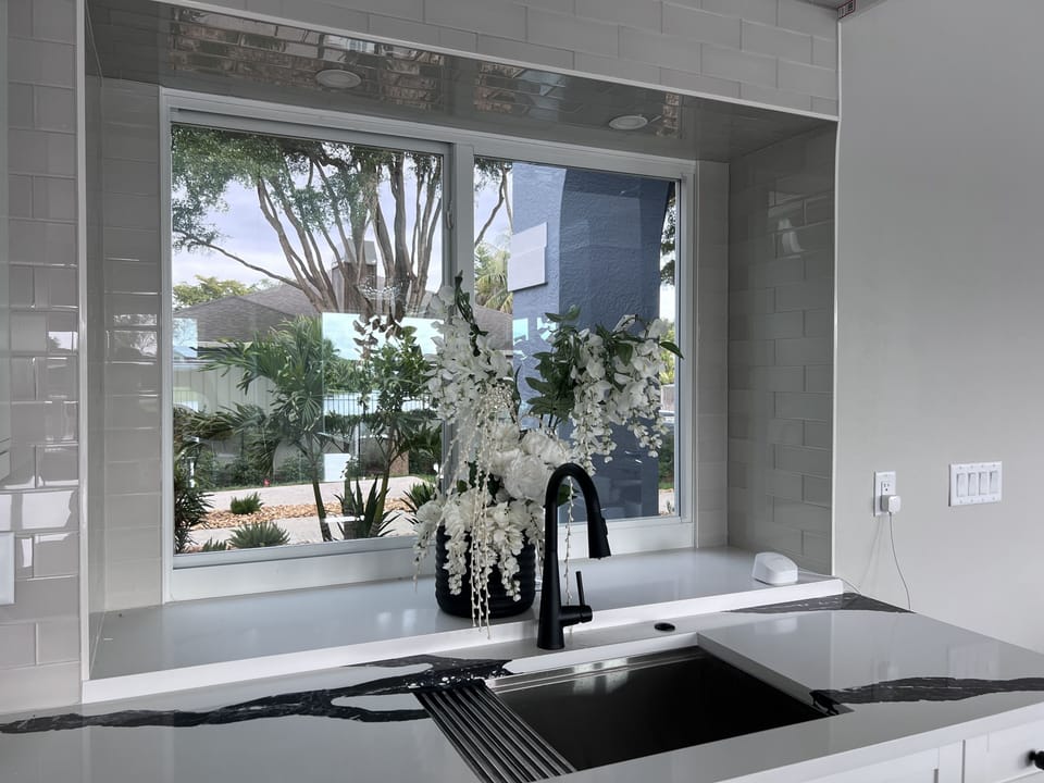A modern kitchen sink with a black faucet, gray countertops, a white backsplash, a window overlooking a garden, and a white floral arrangement in a vase.