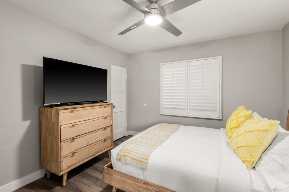 A cozy bedroom with a wood bed frame, matching dresser, and a mounted TV. Soft yellow accents brighten the neutral tones. A ceiling fan provides airflow, and plantation shutters offer light control.