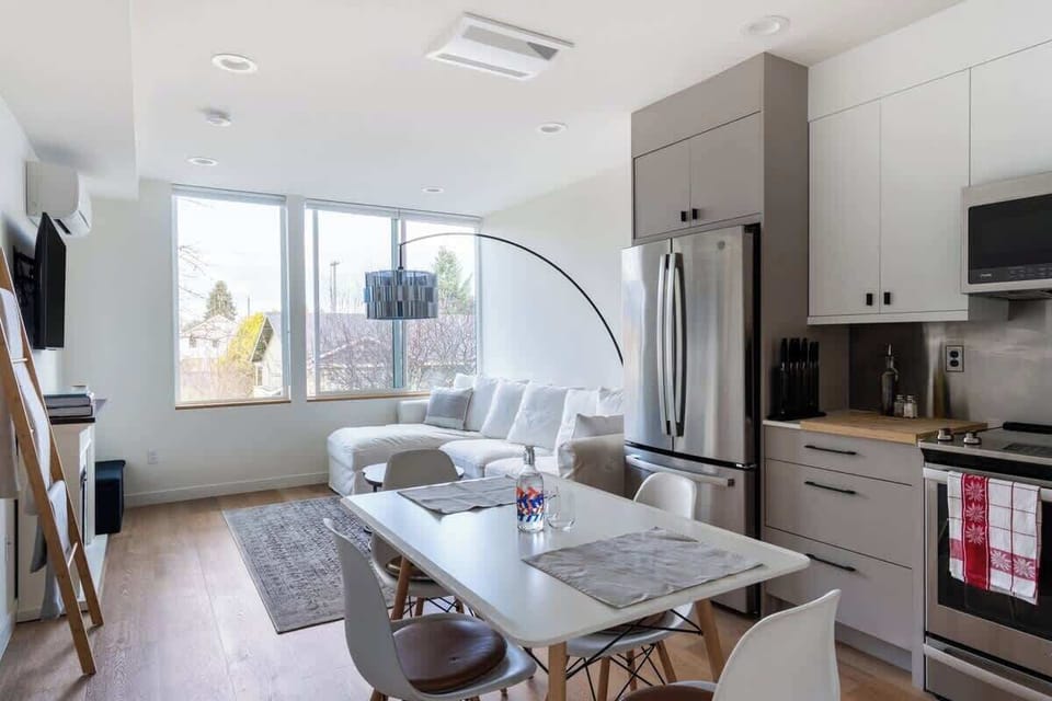 Bright open-concept space with modern kitchen, cozy living area, stylish dining table, and large windows filled with natural light.