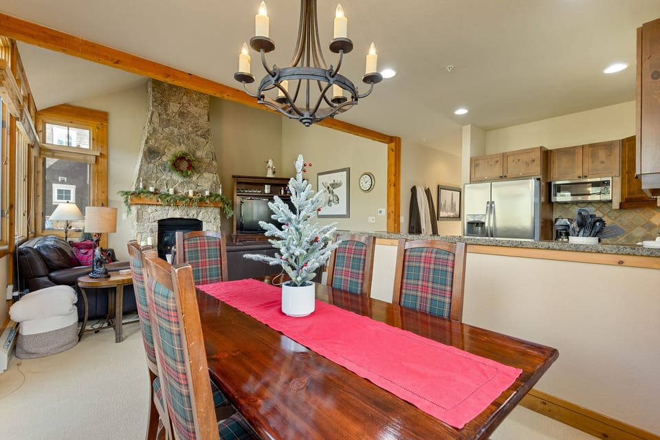A cozy dining room features a wooden table with plaid chairs and a small tree centerpiece. A chandelier hangs above, and a decorated stone fireplace is visible in the background.