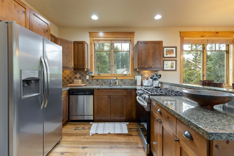 A modern kitchen features stainless steel appliances, wooden cabinets, granite countertops, a tile backsplash, and wooden framed windows providing a view of trees outside.