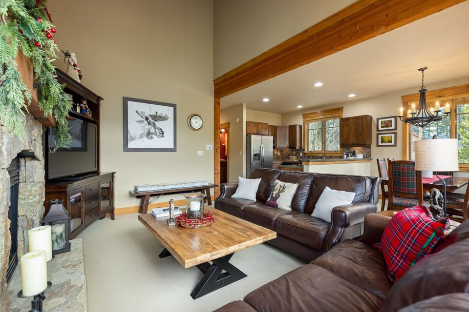 A cozy living room features leather sofas, a wooden coffee table, and a fireplace. Adjacent is an open kitchen and dining area with wooden cabinets and modern appliances. A framed wolf print adorns the wall.