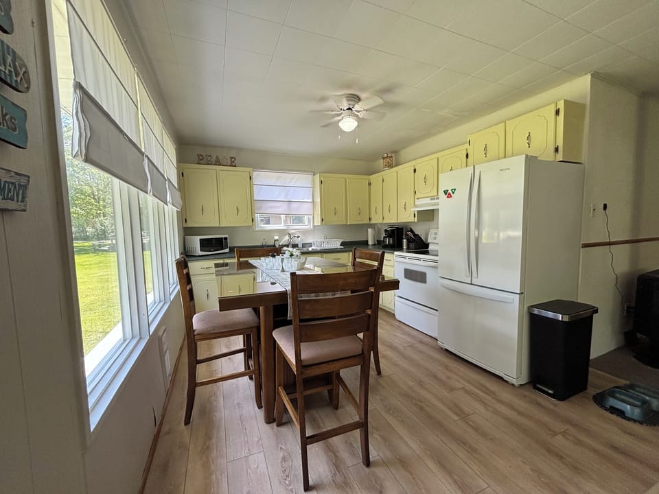 A bright, vintage kitchen with pale yellow cabinets and white countertop. Large windows provide abundant natural light, highlighting the wood laminate flooring. The kitchen is equipped with modern appliances, including a white refrigerator, stove, and microwave, creating a functional and inviting space.