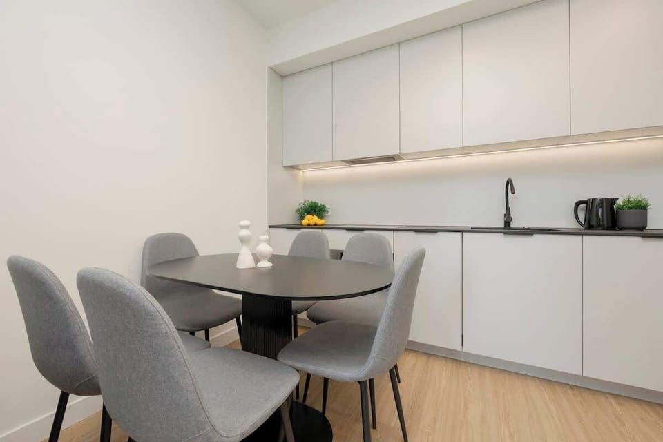 A modern open-plan kitchen and dining area with sleek white cabinets, a black backsplash, and a minimalist dining table with four chairs.

