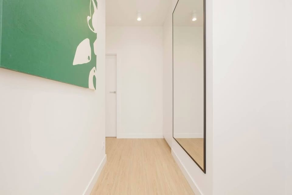 A well-lit hallway with minimalist decor, featuring a full-length mirror and a green abstract painting on the wall.

