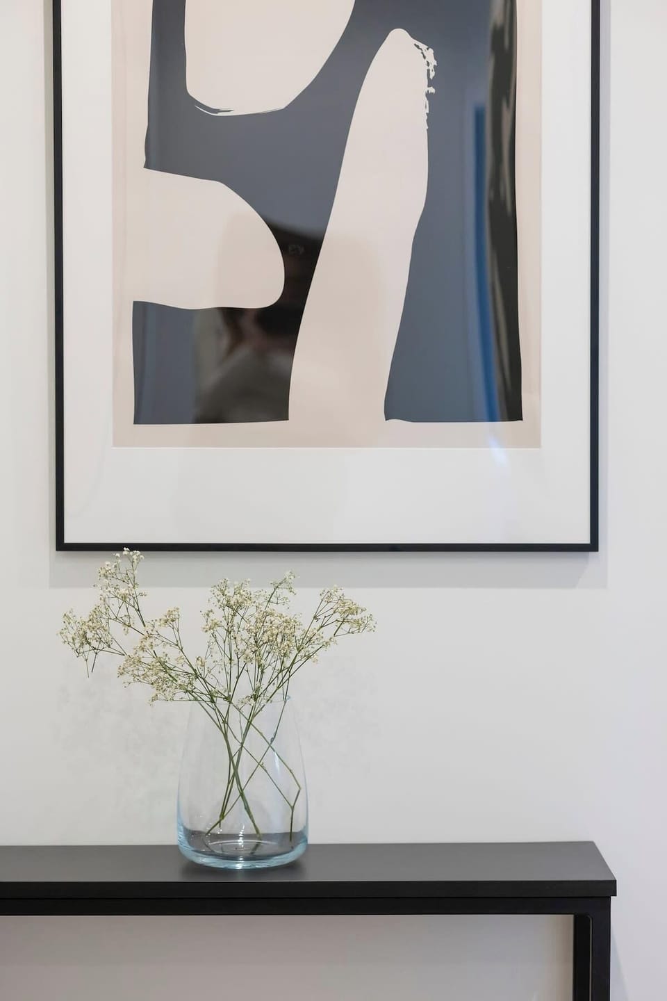 A minimalist wall-mounted console with a framed black-and-white art piece and a sleek white vase with fresh greenery.

