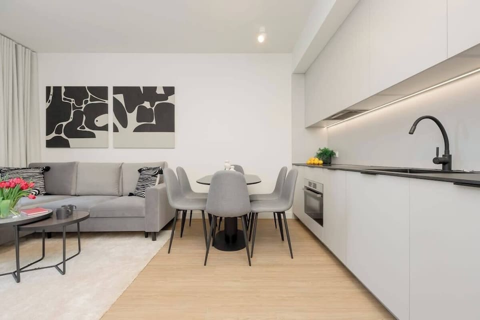 A modern open-plan kitchen and dining area with sleek white cabinets, a black backsplash, and a minimalist dining table with four chairs.

