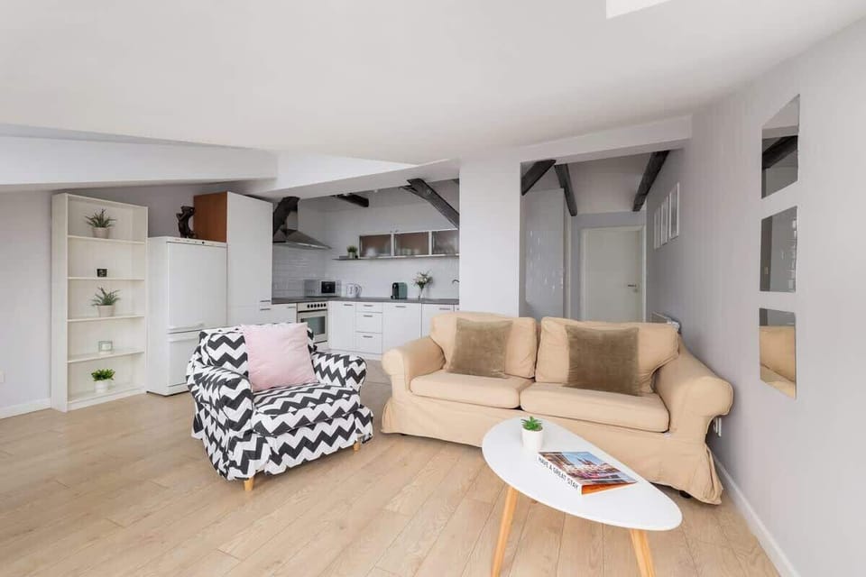 An open living area featuring a beige sofa, a small wooden coffee table, and a patterned armchair, all set against white walls and exposed beams.

