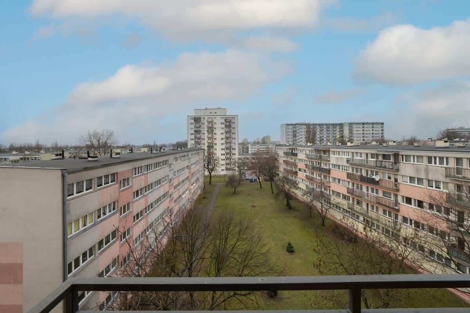 A balcony view overlooking a residential area, showing neighboring apartment buildings and a green courtyard below.







