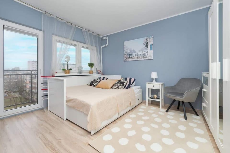 Another angle of the bedroom, showing a large window with sheer curtains, a seating area, and a patterned rug adding comfort to the space.

