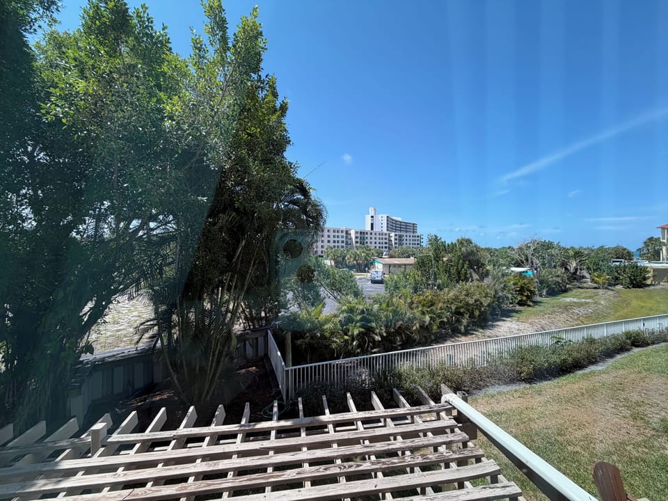 A peaceful riverfront setting featuring lush greenery, mature trees, and well-maintained lawns. The property is situated near the water with urban conveniences visible in the surrounding area.