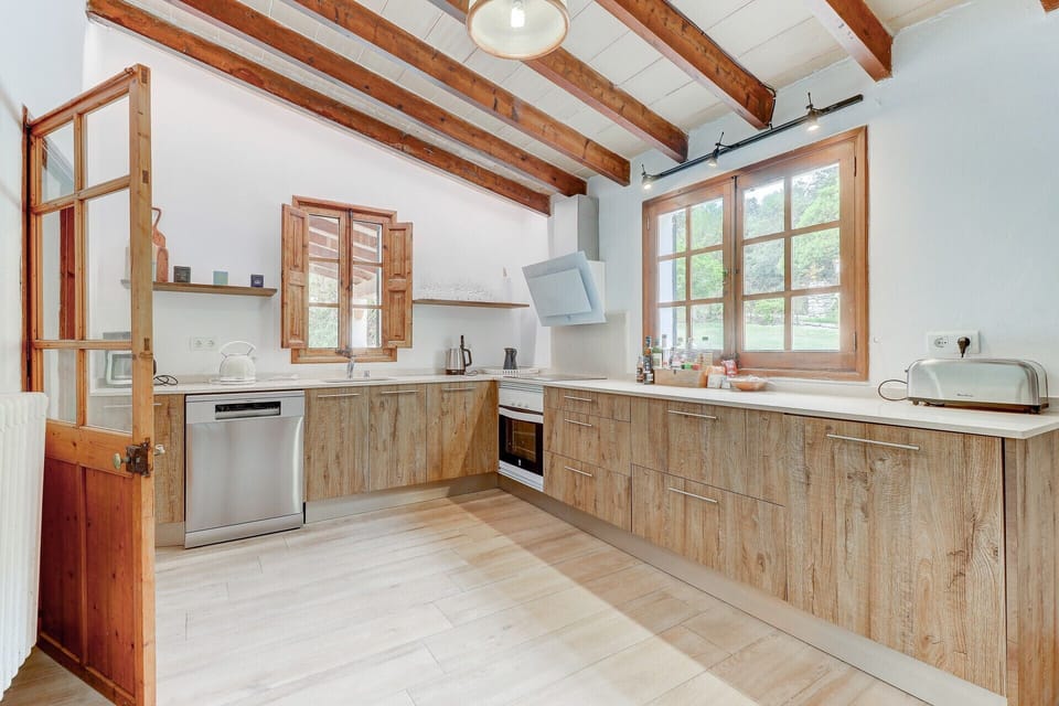 Fully equipped rustic kitchen at Casa Colonya in Pollensa, featuring exposed wooden beams, natural wood furniture, modern appliances, and large windows that provide plenty of natural light and views of the surrounding landscape.