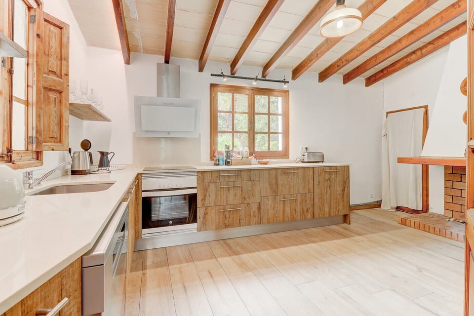 Bright kitchen at Casa Colonya in Pollensa, fully equipped in a rustic-modern style, featuring wooden cabinets, complete appliances and exposed wooden beams. A functional and welcoming space, ideal for preparing meals during a holiday in Mallorca.