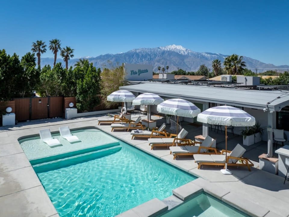 Sunny pool deck with in-water lounge chairs, teak sun loungers, striped cabana umbrellas, and snow-capped mountain views — the desert at its best