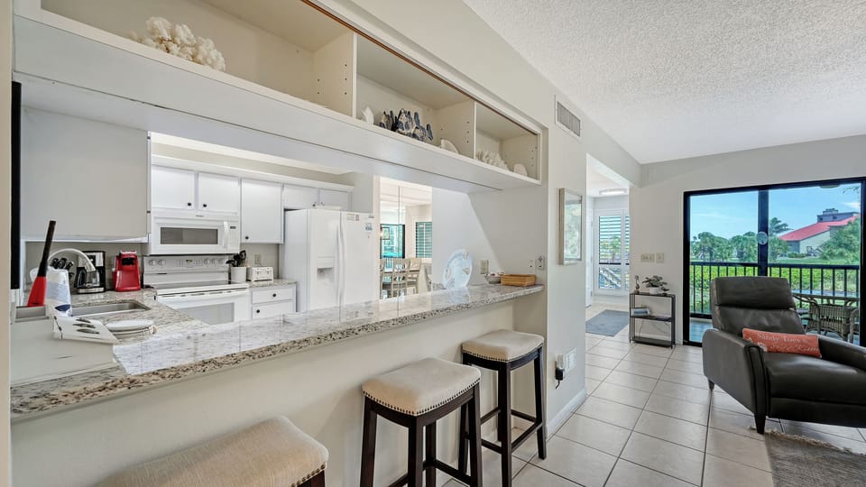 Your open-concept kitchen flows seamlessly into the living area, with granite counters, bar seating, and views that stretch out to a sunny balcony and tree-lined horizon beyond.