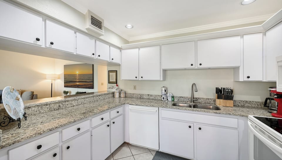 Your fully equipped kitchen features granite countertops, crisp white cabinetry, and everything you need to prepare meals during your stay, with an inviting living area just beyond.