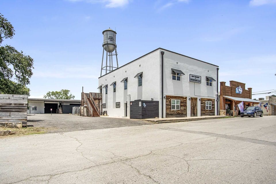 An industrial-style white building featuring rustic wood siding, black metal awnings, and a towering water tank in the background for a unique, small-town aesthetic.