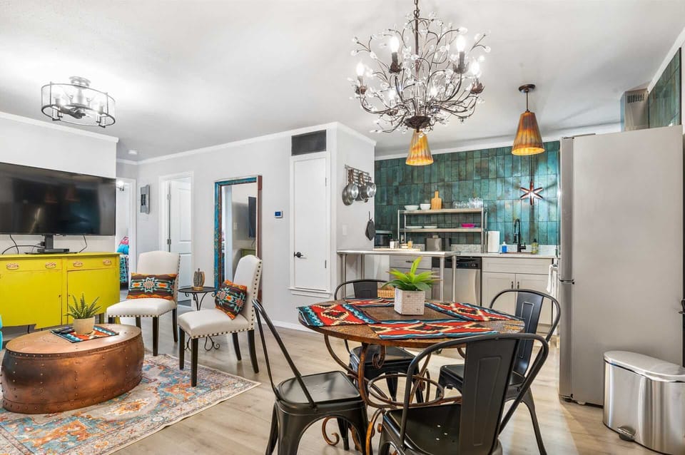 A dynamic, open-concept living and dining area featuring a stunning teal-tiled kitchen backsplash, a copper-toned coffee table, and an ornate crystal chandelier that adds a touch of glamour.