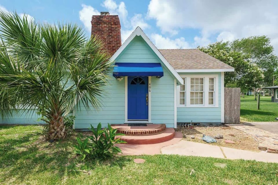  Charming coastal cottage with a bold blue door and tropical landscaping — a warm welcome awaits.