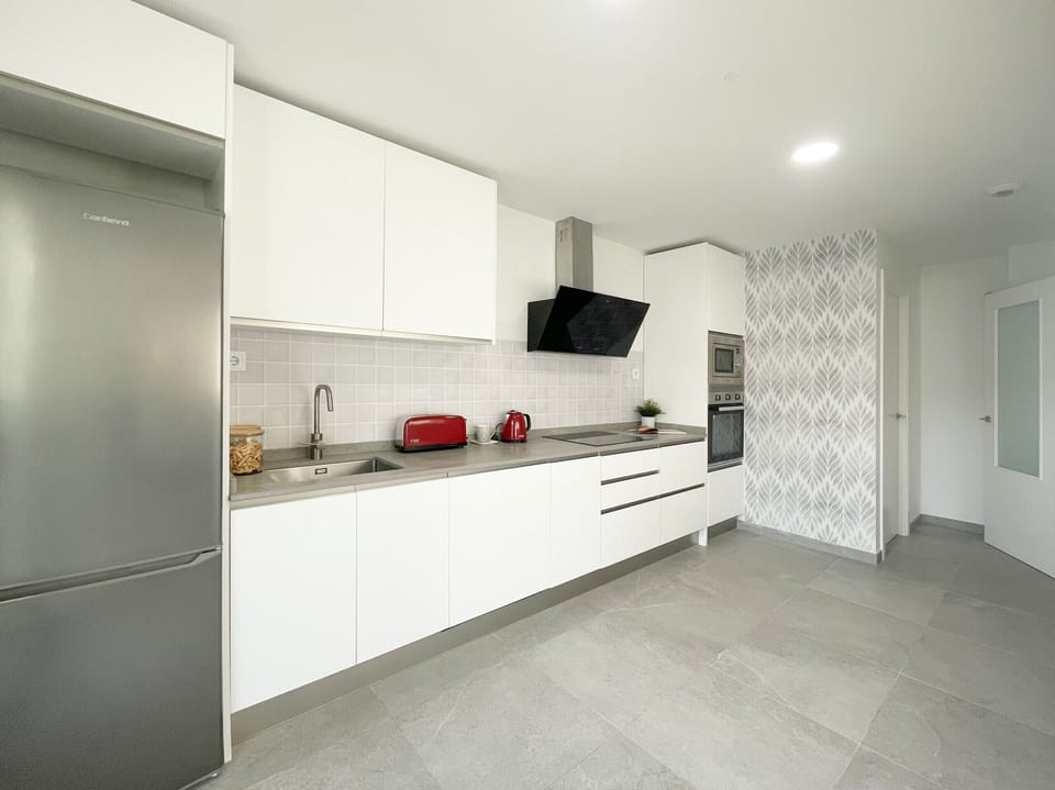 Fully equipped kitchen with a modern and minimalist design, featuring white handleless cabinets, a grey countertop, stainless steel appliances, and elegant porcelain floor tiles. The grey patterned accent wall adds a decorative touch.