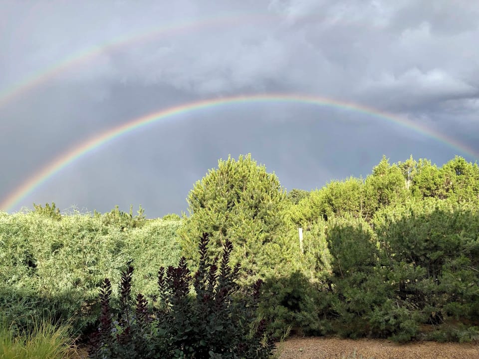 A stunning double rainbow arcs over lush greenery and towering trees in the surrounding natural landscape, creating a magical backdrop for the property.