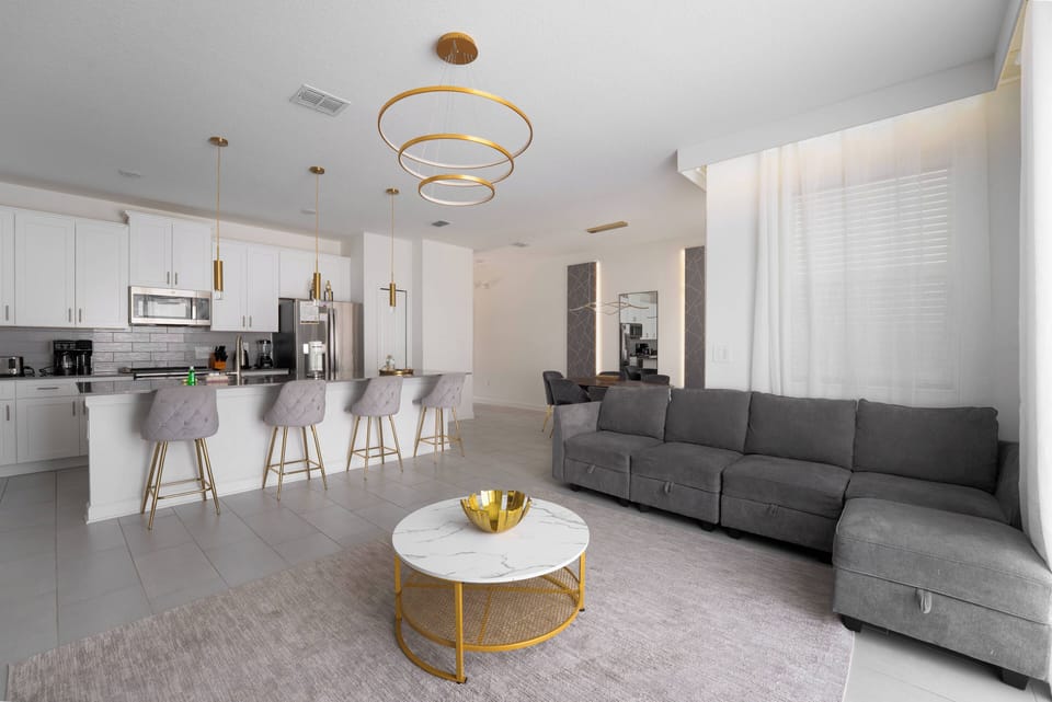 Experience the inviting charm of this spacious living area, featuring a sleek gray sectional, a chic marble coffee table, and stylish bar seating that flows into the modern kitchen, all bathed in warm, natural light.
