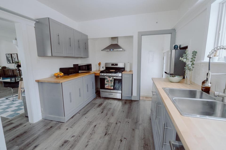 A modern kitchen with grey cabinets, wooden countertops, a stainless steel stove with a hood, a double sink, and various small appliances. The flooring is a dark wood laminate.