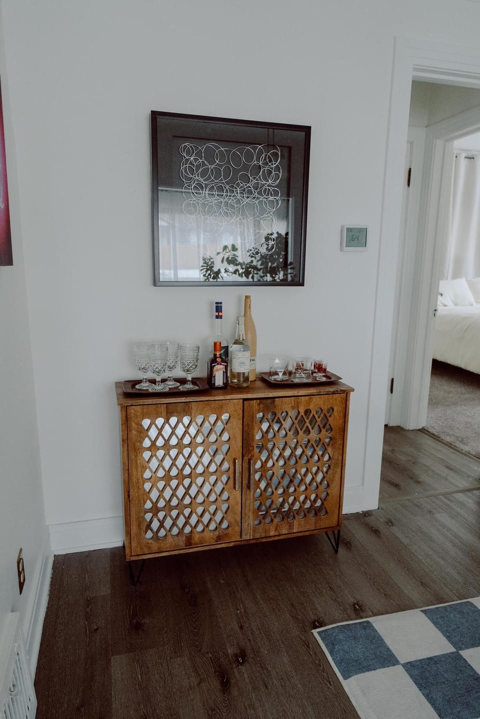 A wooden sideboard in a modern living space holds various bottles and glassware. Above the sideboard, a framed geometric artwork is hung on the white wall. A bedroom is visible in the background.