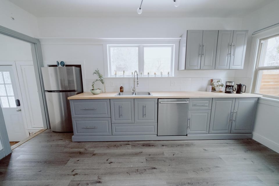 Modern kitchen with gray cabinets, light wood countertops, stainless steel refrigerator and dishwasher, and hardwood floors. Large windows provide natural light. Small plant and coffee maker on counter.